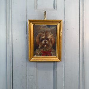 以下が含まれることがあります： 赤いリボンをつけた犬の額入り肖像画。絵画は、金色のフレームに入った、ふわふわの薄茶色と白の犬が特徴です。アートワークは水色のドアに掛けられています。