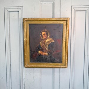 May include: An oil painting in a weathered gold-toned wooden frame depicting a portrait of a person wearing a dark patterned garment, a light-colored shawl, and a bonnet. The painting shows visible craquelure across the surface of the canvas.