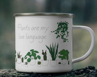 May include: White enamel mug with a silver rim and handle. The mug features the text "Plants are my love language" and a line of green plant illustrations. The mug is sitting on a tree trunk.