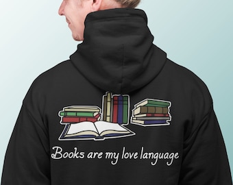 Puede incluir: Sudadera con capucha negra con un gráfico de libros apilados y un libro abierto. El texto "Books are my love language" está impreso en blanco debajo. La sudadera es usada por una persona, vista de espaldas.
