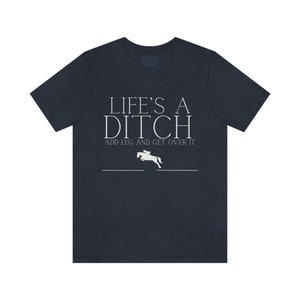 Puede incluir: Una camiseta azul oscuro con el texto "Life's a Ditch Add Leg and Get Over It" y una silueta blanca de un caballo saltando una valla.