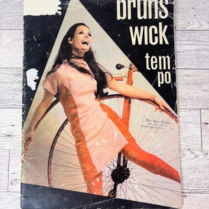 May include: Vintage magazine cover featuring a woman in a pink dress and orange tights, posing with a bicycle. The cover has the words "Brunswick Tempo" in white text. The background is black and white with a geometric design.