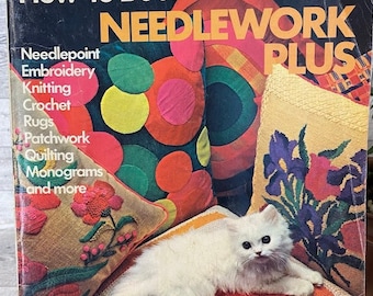 May include: A vintage book titled "Simplicity's How-to Book Needlework Plus" featuring a white cat lying on a colourful knitted cushion. The book cover includes a list of needlework projects such as needlepoint, embroidery, knitting, crochet, rugs, patchwork, quilting, monograms, and more. The price is £1.00.