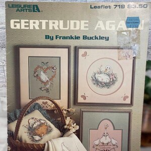 Op de afbeelding: Brochure met borduurpatronen met een verzameling ontwerpen met een landelijk thema. De ontwerpen omvatten een gans in een nest, een gans met een mand vol eieren en een gans met een welkomstbord. De brochure heeft de titel "Gertrude Again" van Frankie Buckley.