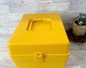 Puede incluir: Una caja de almacenamiento rectangular de color amarillo brillante con un asa en la parte superior y un cierre de pestillo. La caja está hecha de plástico y tiene un acabado ligeramente brillante. La caja está sobre una superficie de madera gris claro y blanca.
