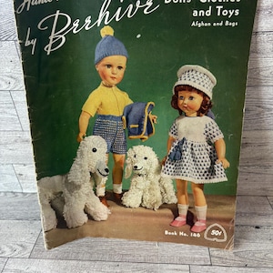 Può includere: Una copertina di libro vintage che presenta un libro di modelli di vestiti e giocattoli per bambole lavorati a maglia a mano. La copertina presenta due bambole, un bambino e una bambina, con i loro abiti lavorati a maglia e due cani lavorati a maglia bianchi. Il titolo del libro è "Hand Knits by Barhive Dolls' Clothes and Toys Afghan and Bags" e il numero del libro è 146.