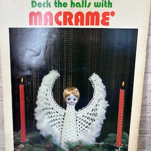May include: A vintage book cover with the text "Deck the halls with MACRAME". The image features a macrame angel with white wings, a doll face, and two red candles on either side. The angel is surrounded by greenery.