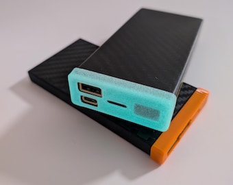 May include: Two black power banks with a carbon fibre pattern. The top power bank has a light blue protective cover with two gold USB ports. The bottom power bank has an orange protective cover.