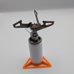 Puede incluir: Una estufa de camping compacta con un cartucho de combustible blanco y naranja. La estufa tiene un cabezal de quemador de metal oscuro con brazos plegables y una base triangular naranja. Ideal para cocinar al aire libre.