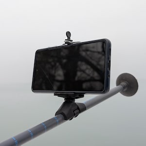 Puede incluir: Un teléfono inteligente negro conectado a una pértiga telescópica gris con una empuñadura de goma negra. El teléfono está montado en un soporte de teléfono negro con un pequeño botón negro en la parte superior.