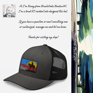 May include: A gray and black trucker hat with a colorful embroidered patch featuring a silhouette of a person riding a horse. The patch has a blue, yellow, red, and white color scheme. The text "Brushstroke BoulevArt" is visible on the right side of the image.