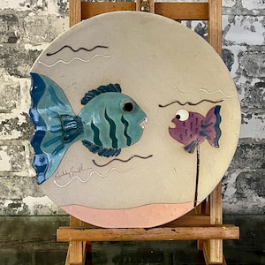 May include: A round ceramic art piece featuring two stylized fish. One fish is teal with dark blue accents, the other is purple. The background is beige with wavy lines and a pink base. Signed "Kelly Smith".