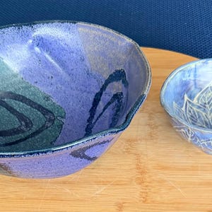 Może przedstawiać: Dwie ceramiczne miski w odcieniach niebieskiego i fioletowego. Większa miska ma nieregularny kształt i ciemnoniebieskie wiry. Mniejsza miska ma biały kwiatowy wzór. Obie miski znajdują się na drewnianej powierzchni.