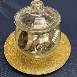 May include: A clear glass jar with a lid, filled with metal cookie cutters, sits on a round, gold glitter-covered plate. The jar is round with a small handle on the lid. The plate is a solid gold color and reflects light.