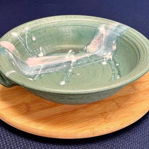 Peut inclure: Un bol en céramique vert fait à la main avec un design unique. Le bol a une forme ronde avec un bord légèrement évasé et deux petites poignées. Il présente des accents blancs et roses et repose sur une assiette en bois ronde.