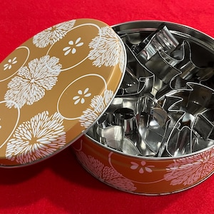 May include: A round, open tin with a floral design on the lid and rim, containing various metal cookie cutters. The lid is off, revealing the cutters inside. The tin is gold with white floral patterns.