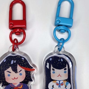 Puede incluir: Dos llaveros de acrílico transparente con purpurina incrustada en el plástico. Los llaveros representan personajes de anime con cabello negro y atuendos diferentes. Un llavero tiene un clip rojo y azul, el otro tiene un clip azul.
