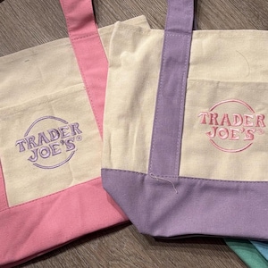 May include: Two canvas tote bags, one pink and one lilac, with cream-coloured centres and pockets. Each bag features the embroidered "Trader Joe's" logo in a circular design. The bags have matching coloured straps and bottom panels.