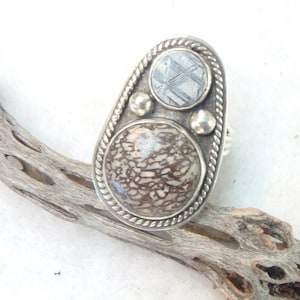May include: A silver ring with an oval face featuring two circular stones and two small silver beads. The larger stone is a light brown color with a patterned design. The smaller stone is a light gray color with a linear pattern.