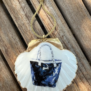 以下が含まれることがあります： 金色の縁と金のロープハンガーが付いた白い貝殻の飾り。貝殻には青と黒のトートバッグのデザインが施されています。背景は木製の表面です。