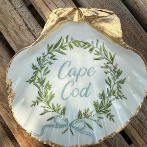 Puede incluir: Una concha blanca con borde dorado y la inscripción "Cape Cod" en azul claro. La concha está decorada con una corona floral verde y morada y una cinta azul.