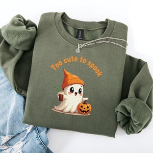 May include: An olive green sweatshirt with the phrase "Too cute to spook" in orange. The graphic features a cartoon ghost wearing an orange beanie and holding a coffee, with a jack-o'-lantern. A silver necklace is draped on the shirt.