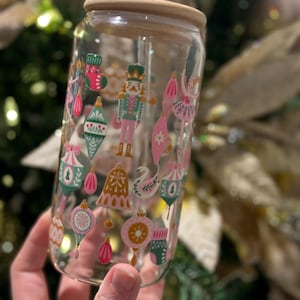 Op de afbeelding: Een heldere glazen beker met een houten deksel en een rietje. De beker is versierd met kleurrijke kerstthema-illustraties, waaronder ornamenten, een notenkraker en een ballerina. Het ontwerp heeft roze, groene en gouden accenten.