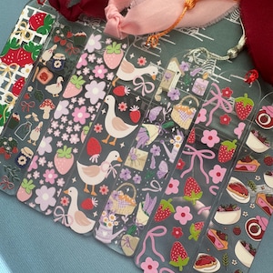 May include: A collection of clear acrylic bookmarks with various designs. The bookmarks feature illustrations of strawberries, flowers, ducks, and bows. Each bookmark has a different design and color scheme, with pink, red, and green accents. Some have tassels.