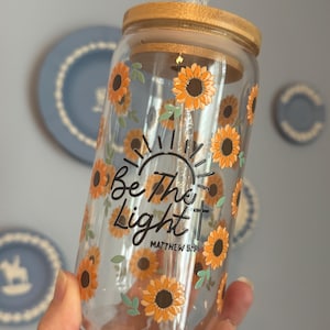 Puede incluir: Un vaso de vidrio transparente con tapa de bambú, adornado con girasoles naranjas y el texto "Be The Light" con un diseño de cruz y sol. También se incluye el texto "Matthew 5:16". El vaso tiene una pajita.
