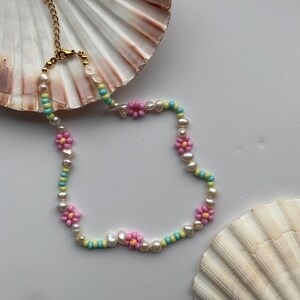 May include: A delicate beaded necklace with pastel pink, yellow, and blue beads and small pink flower charms. The necklace is displayed on a white background with a seashell.
