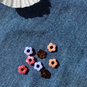 Pode incluir: Conjunto de contas de resina coloridas em forma de flor em rosa, roxo e marrom. As contas estão espalhadas sobre um fundo de jeans azul.