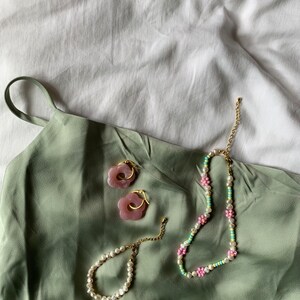 May include: A set of pink flower-shaped earrings, a pearl bracelet, and a beaded necklace with pink and green flowers. The jewelry is on a green fabric background.