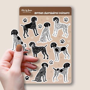 Peut inclure: Planche d'autocollants avec des illustrations de braques allemands à poil court dans diverses poses. Les chiens sont en noir et blanc, avec des empreintes de pattes et des cœurs. Le texte "German shorthaired pointers" est visible.