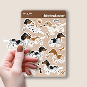 May include: A sheet of stickers featuring piebald dachshund dogs in various poses. The stickers are on a brown background with white paw prints, bones, and stars. The text "piebald dachshunds" is at the top of the sheet.