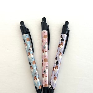 May include: Three black pens with different coloured patterned designs featuring dogs. The pens have black caps and are arranged in a triangular shape.