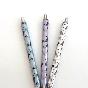 May include: Three pens with different colored bodies and a black and white pattern of dogs and bones. The pens are all silver and have a black ink cartridge.