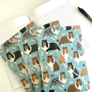 May include: A set of four blue notebooks with a pattern of colorful dogs on the cover. The notebooks are stacked on top of each other.