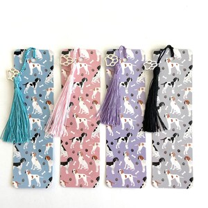 May include: Four bookmarks with a paw print charm and a tassel. Each bookmark features a different color background with a repeating pattern of white and brown dogs.