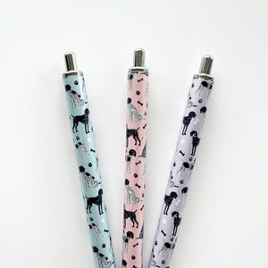 May include: Three pens with dog-themed patterns. One pen is light blue with black dog silhouettes and white spotted dogs. Another is pink with black dogs and white spotted dogs. The third is lavender with black dogs and white spotted dogs.