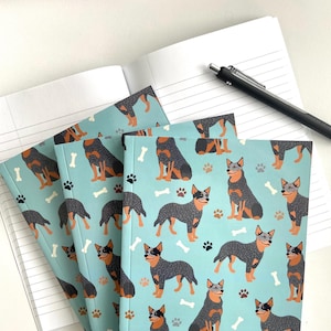 May include: A stack of four blue notebooks with a pattern of Australian Shepherd dogs and bones. The notebooks have white lined pages and a black pen is lying on top of the stack.