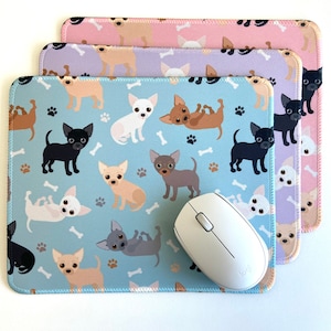 May include: A set of three mouse pads featuring a repeating pattern of cartoon chihuahua dogs in various poses. The mouse pads are in shades of blue, purple, and pink. A white computer mouse is on top of the blue mouse pad.