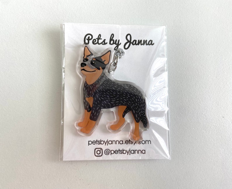 May include: A black and white Australian Cattle Dog keychain with a silver key ring. The keychain is made of clear acrylic and has a cartoon-style illustration of the dog. The text "Pets by Janna" is printed on the card behind the keychain. The text "petsbyjanna.etsy.com" and "@petsbyjanna" are also printed on the card.