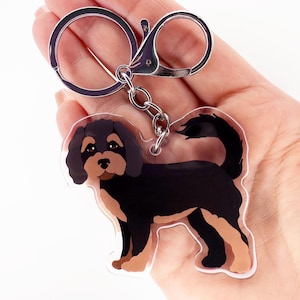 May include: A clear acrylic keychain featuring a cartoon illustration of a Cavalier King Charles Spaniel dog. The dog has black and tan markings and a curly tail. The keychain has a silver-colored clasp and key ring.