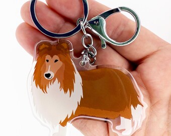 May include: A clear acrylic keychain featuring a stylized illustration of a Collie dog. The dog has brown and white fur with a silver-colored metal keyring and clasp.