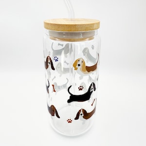 May include: A clear glass tumbler with a bamboo lid and a clear straw. The tumbler is decorated with colorful illustrations of basset hounds, paw prints, and bones. The design is repeated around the entire glass.