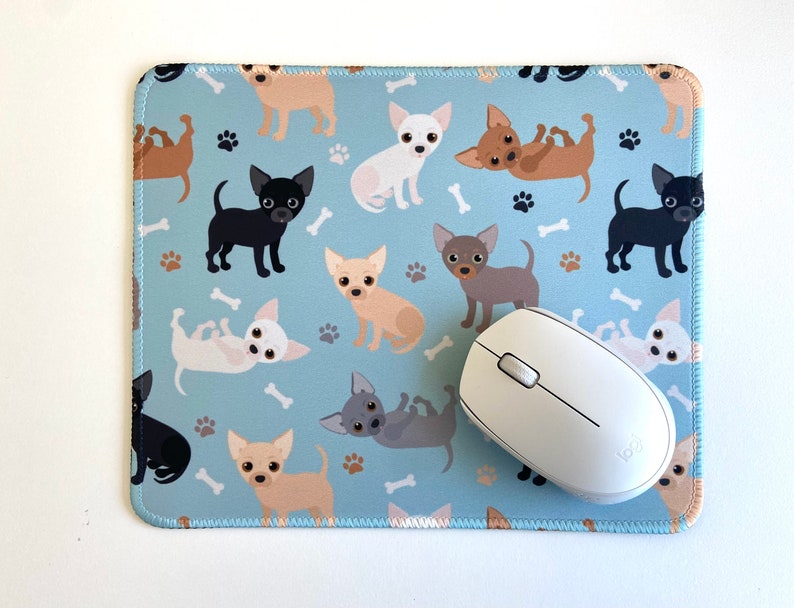 May include: A blue mouse pad with a repeating pattern of white, brown, and black chihuahua dogs. A white wireless computer mouse is on top of the mouse pad.