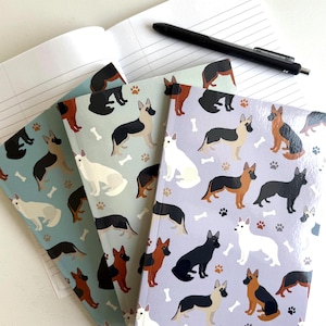 May include: Three notebooks with different colored covers featuring a repeating pattern of German Shepherd dogs in various poses. The covers are blue, green, and purple. The notebooks are stacked on top of each other.