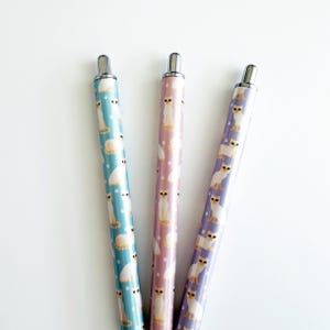 May include: Three retractable ballpoint pens featuring a repeating pattern of white cats with brown ears and tails. The pens have light blue, light pink, and light purple barrels, each topped with a silver-colored metal click mechanism and clip.