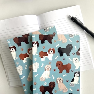 May include: Two blue notebooks with a pattern of brown, black and white dogs on a light blue background. The notebooks are open and a black pen is lying on top of the notebook.