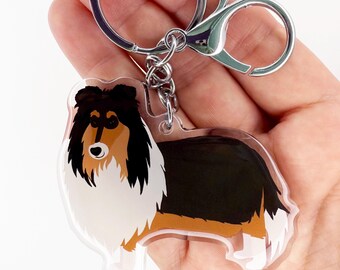 May include: A clear acrylic keychain featuring a cartoon-style Collie dog. The dog has a black, brown, and white coat. The keychain includes a silver-colored split ring and a lobster clasp for easy attachment.